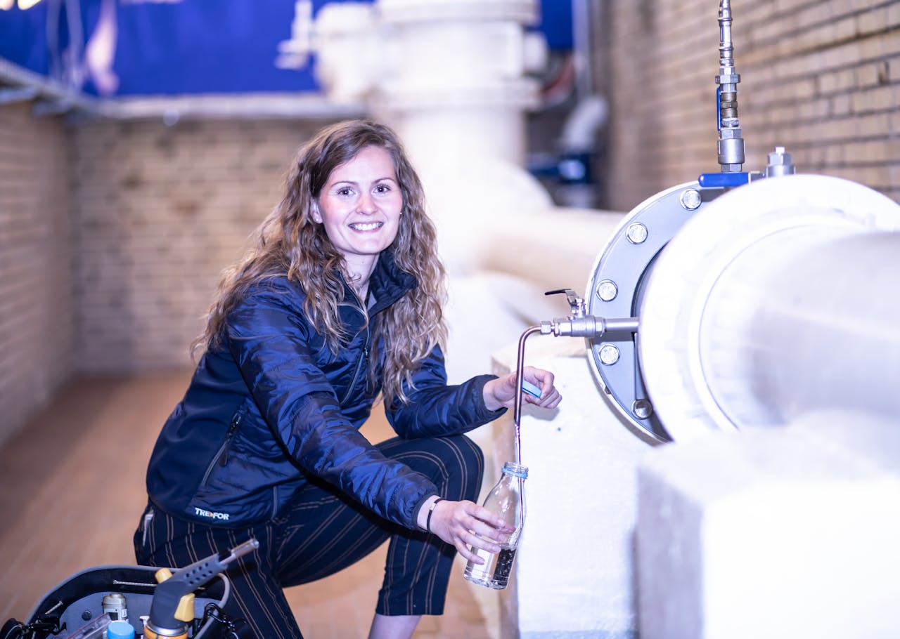 photo of a smiling woman drawing some water with a bottle near a large pipe.